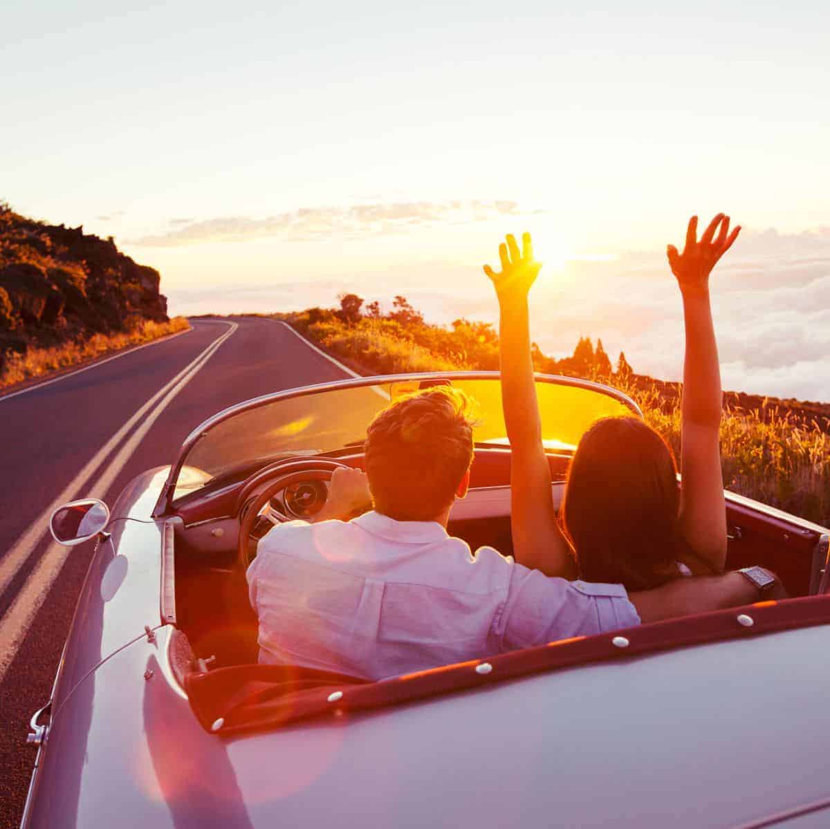 A couple in a small convertible on a winding road at sunset.